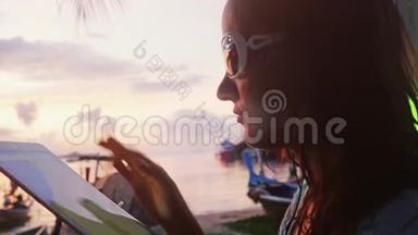 日落时分，在岸上的户外咖啡馆里戴着太阳镜的年轻女子使用平板电脑和喝酒。 假期